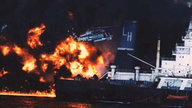 Des flammes et une épaisse fumée noire s'élèvent d'un navire en pleine nuit, tandis qu'un hélicoptère militaire survole les environs. Les flammes brûlent à la surface de la mer.